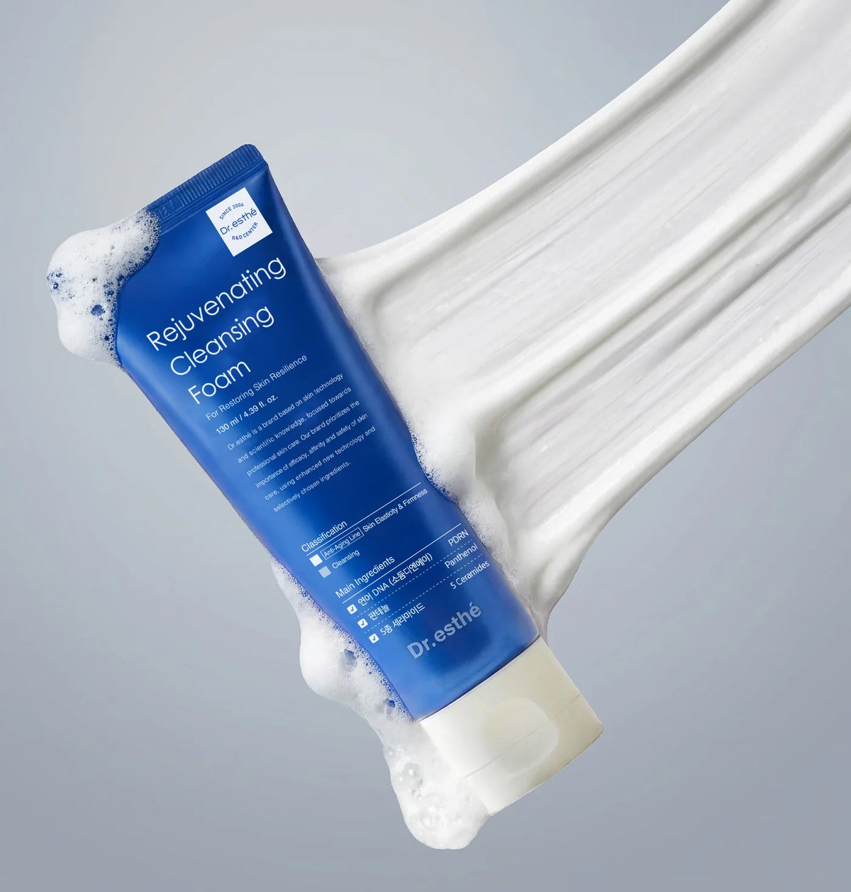A blue tube of Dr. Eustine Rejuvenating Cleansing Foam with white foam, shown against a clean gray background with a dynamic streak of thick white foam behind it.